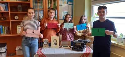 Uczniowie trzymają kartki z cytatami W. Szymborskiej. Przed nimi, na stole, stoi maszyna do pisania, radio, zegar i portret Szymborskiej. 