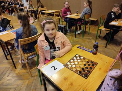 Dziewczynka z przypiętym numerem trzy siedzi przy stoliku oznaczonym cyfrą dwa, na którym leżą warcaby