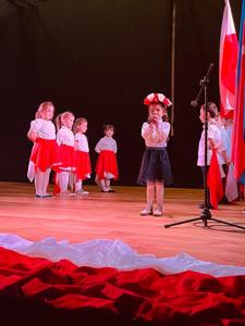 dziewczynka ubrana na biało czarno z wiankiem biało czerwonych kwiatów na głowie  na środku sceny trzyma mikrofon przy buzi. 