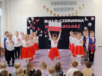  Na środku sali stoi dziewczynka w białej bluzeczce i czerwonej spódniczce trzymająca nad swoją głową flagę Polski