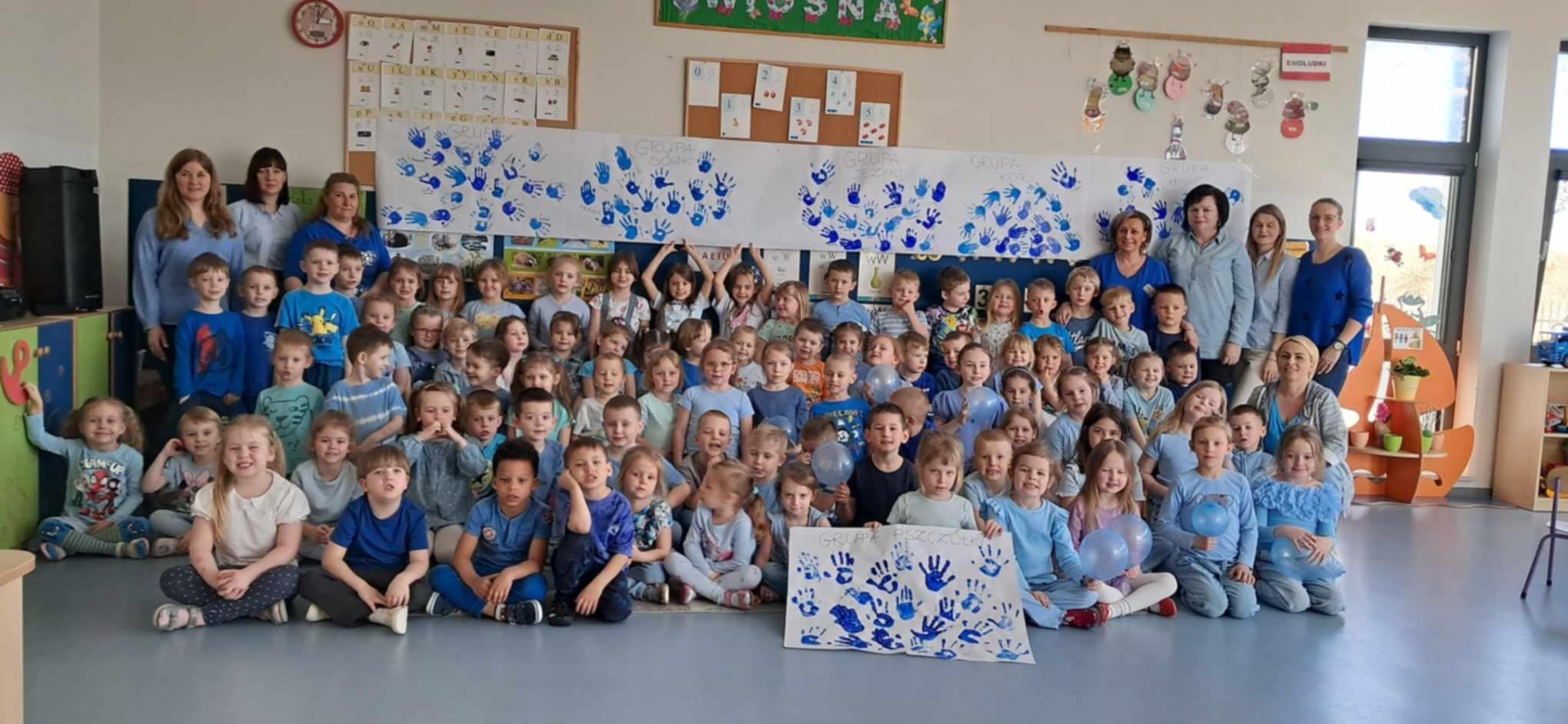 Grupa przedszkolaków ubrana na niebiesko stoi na tle plakatu przedstawiającego odbite w niebieskiej farbie ręce przedszkolaków jako symbol hasła TRZYMAMY Z WAMI MOTYLAMI