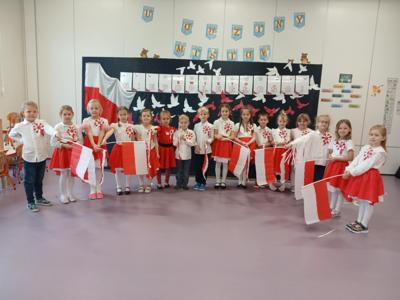 Grupa dzieci w biało czerwonych strojach stojąca na tle dekoracji i napisu Dzień odzyskania Niepodległości 