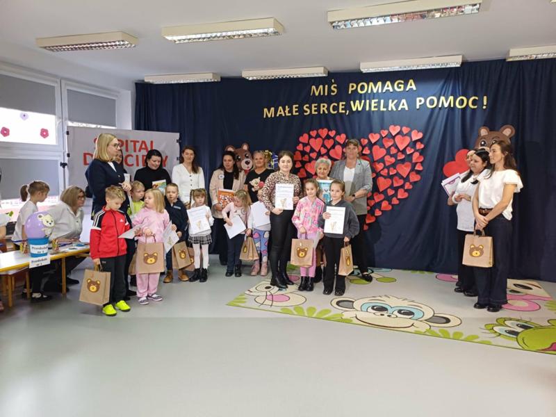 Grupa dzieci wraz z Paniami nauczycielkami stojąca na tle granatowego materiału na którym widnieje wielkie serce zrobione z małych serc i napis Miś pomaga, wielkie serce wielką pomoc 