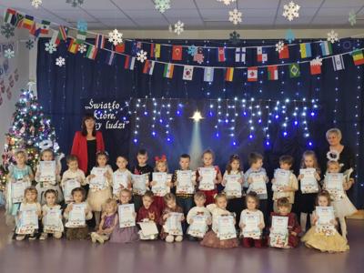 Grupa przedszkolaków stojąca z dyplomami na niebieskim tle. Po bokach stojące Pani dyrektor i Pani organizator konkursu