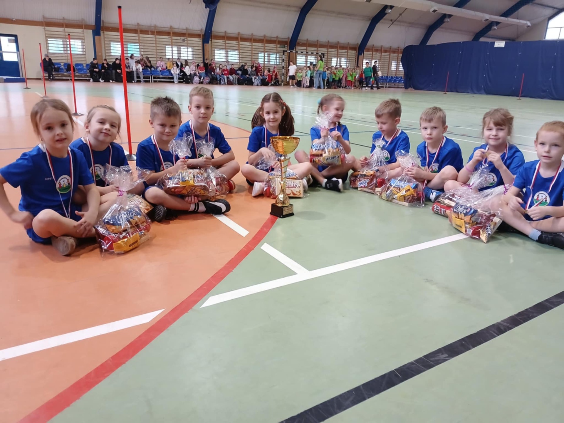 Grupa dzieci biorących udział w zawodach siedzi w półokręgu a przed nimi stoi puchar