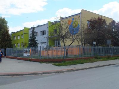 Pomarańczowo, biało, zielony budynek przedszkola nr 2 w Koluszkach umiejscowiony wśród drzew.