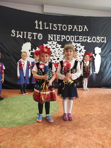 Dzieci w strojach krakowskich stoją na tle granatowego materiału z napisem 11Listopada - Święto Niepodległości. Na środku stoją dwie dziewczynki, krakowianki trzymające mikrofony. dziewczynki śpiewają