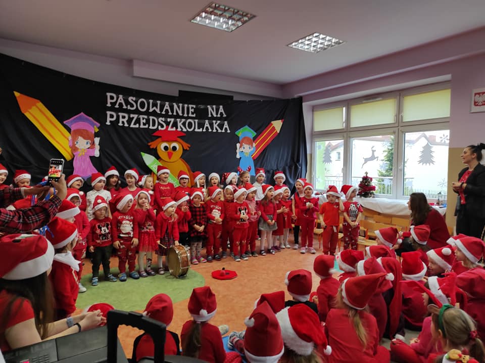 dzieci ubrane na czerwono w czapkach Mikołaja na tle granatowego materiału z napisem Pasowanie na przedszkolaka