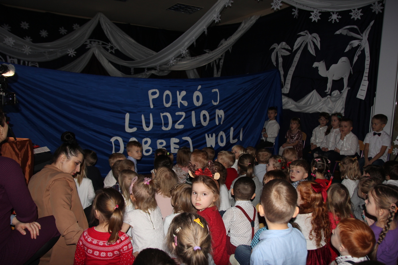 Przedszkolaki siedzące tyłem, zwrócone w kierunku granatowego materiału z białym napisem "Pokój ludziom dobrej woli"