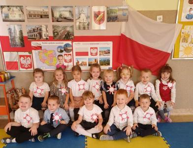 Dzieci w odświętnych  strojach z kotylionami w barwach narodowych siedzą na tle gazetki patriotycznej