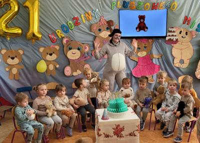 Grupa dzieci oraz nauczycielka w stroju misia na tle niebieskiego materiału z napisem Urodziny pluszowego misia