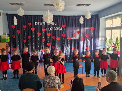 grupa dzieci ubrana w czarno- czerwone stroje, stojąca na tle granatowego materiału, do którego przyczepione są kwiatki z serduszek. Dzieci występują przed dziadkami