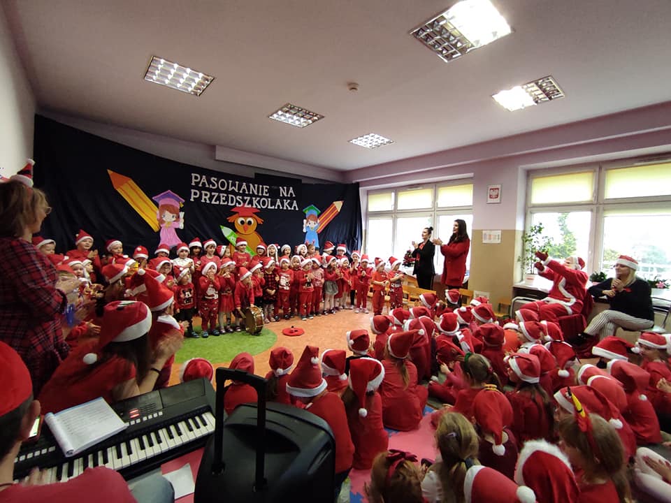 Dzieci w przedszkolu ubrane w czerwone stroje i mikołajowe czapki. W tle granatowy materiał z napisem pasowanie na przedszkolaka oraz dziewczynka i chłopiec z kredkami