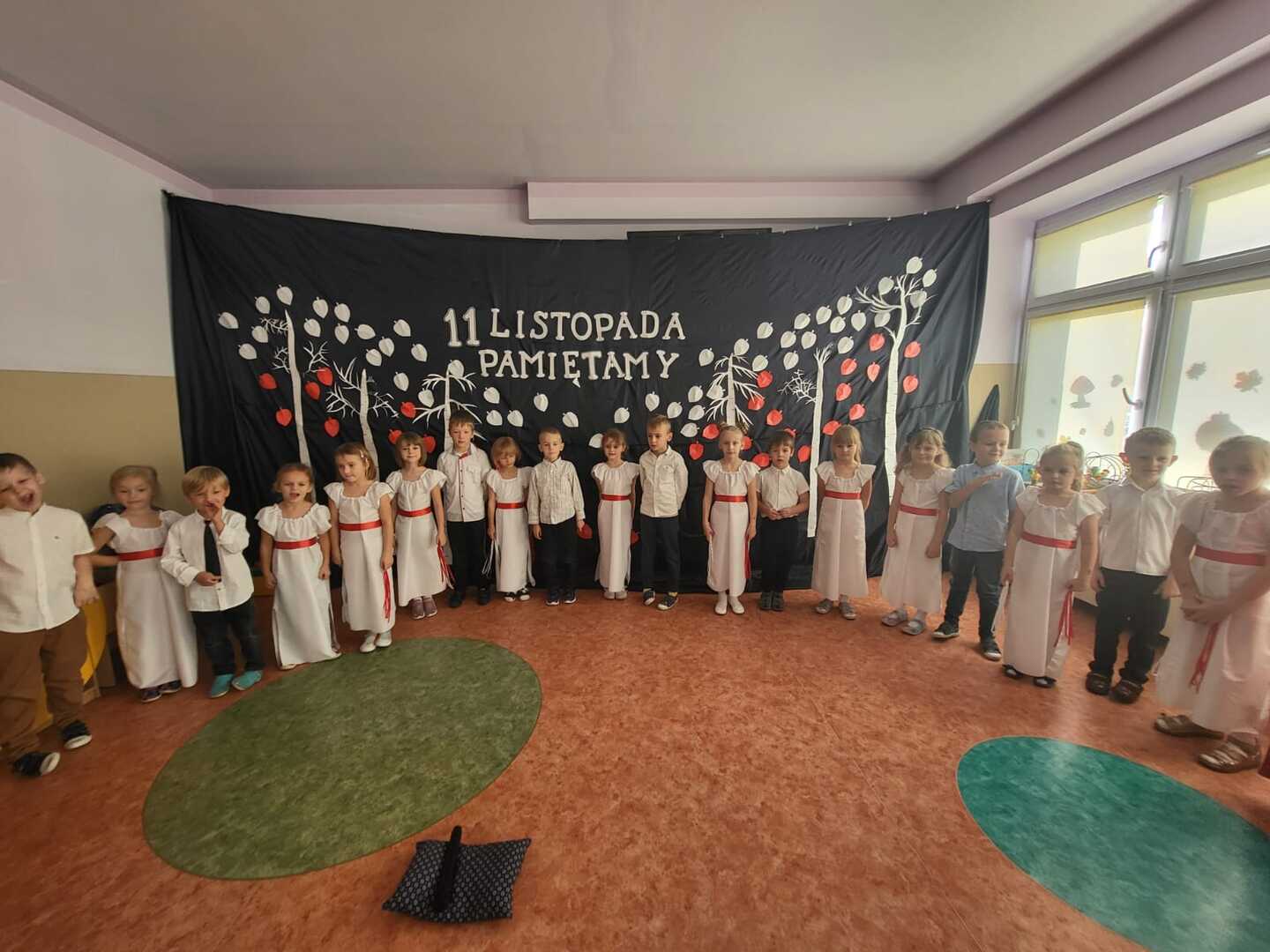 Dziewczynki w barwach narodowych, chłopcy ubrani odświętnie, na tle patriotycznej dekoracji z napisem 11 Listopada- Pamiętamy