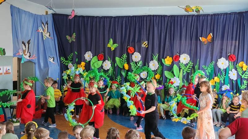 grupa tańczących dzieci ubrana w kolorowe wiosenne stroje -  biedronek, pszczółek kwiatków.  Dzieci tańczą na tle wiosennej dekoracji 