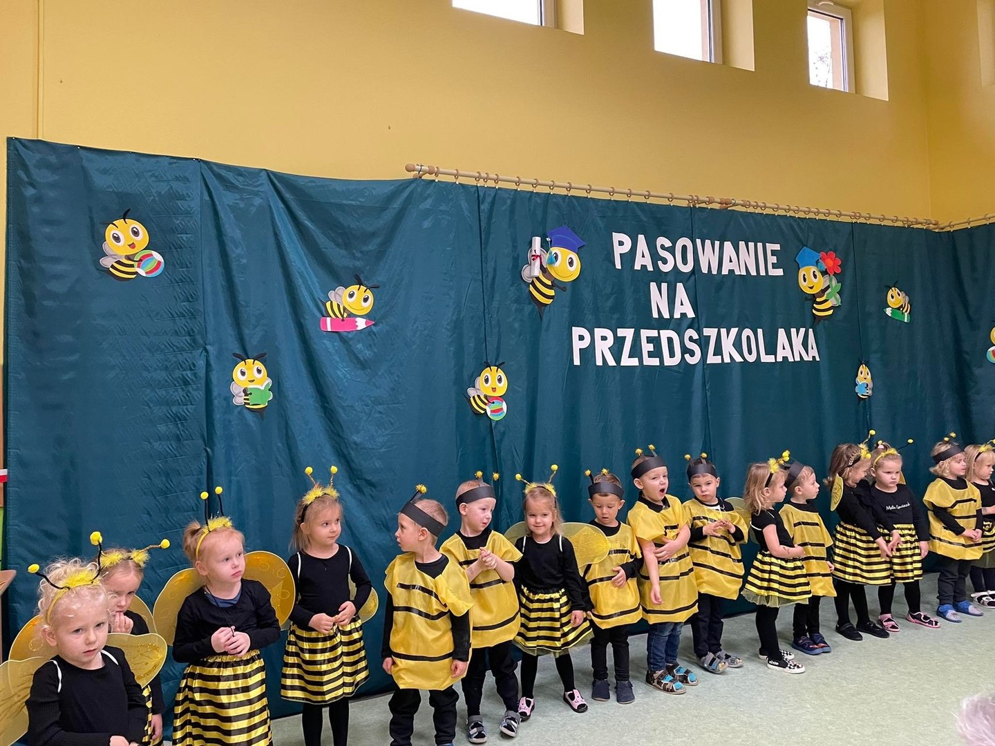 Dzieci ubrane w stroje pszczółek podczas swojego występu na tle dekoracji - pszczółki i napis "Pasowanie na przedszkolaka".