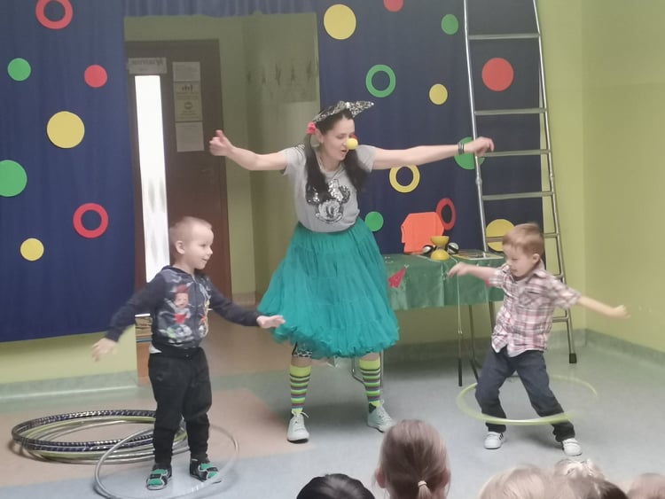 Dwoje dzieci kręci hula hop,pomiędzy nimi stoi artysta cyrkowy.