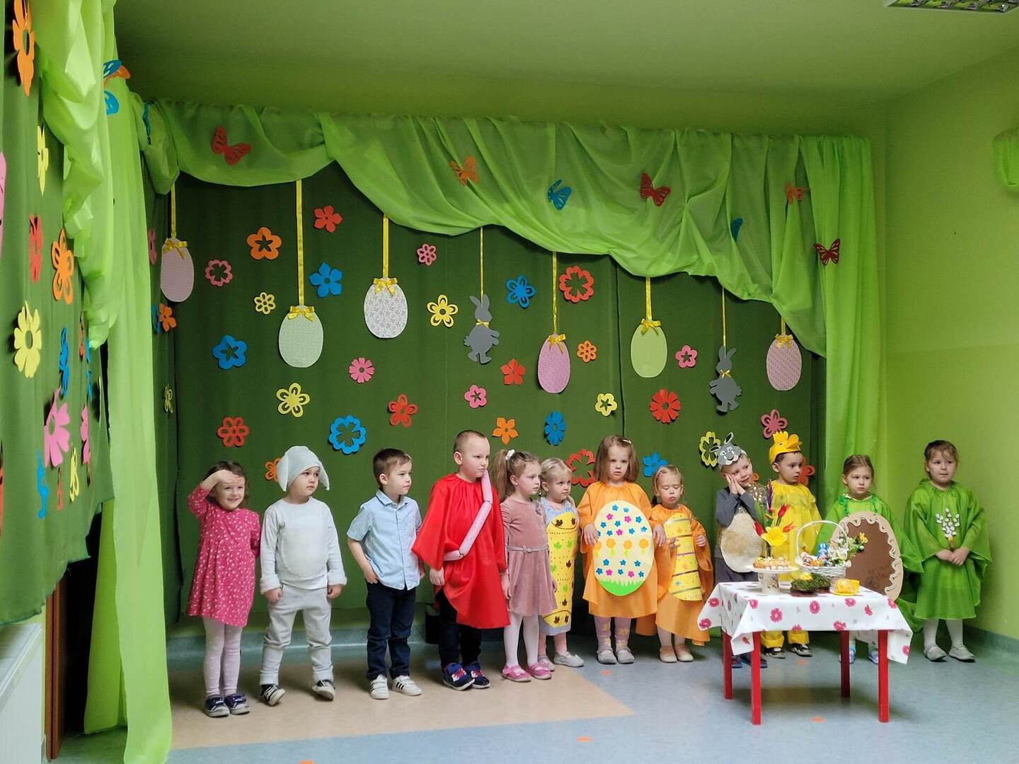 Dzieci w przebraniach stoją w szeregu, przed nimi stolik z koszyczkiem wielkanocnym.
