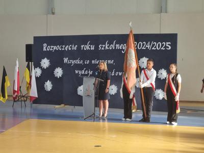 Pani Dyrektorka stoi przy mównicy. Obok mównicy stoi poczet sztandarowy. W poczcie 2 dziewczynki i jeden chłopiec. Chłopiec trzyma sztandar. Za mównicą granatowa tablica z napisem: Rozpoczęcie Roku Szkolnego 2024/25. Wszystko co nieznane wydaje się cudowne.