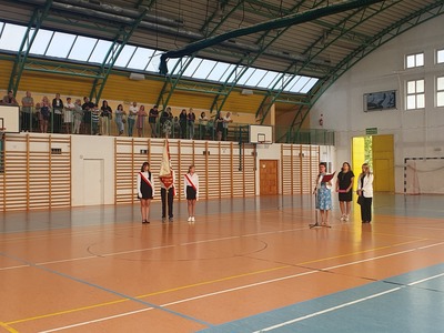 Rozpoczęcie roku szkolnego na hali sportowej. Na pierwszym planie dwie uczennice oraz uczeń trzymający sztandar szkoły. Dyrektor szkoły przemawiająca do mikrofonu. Na drugim planie grupa rodziców stojących na trybunie.