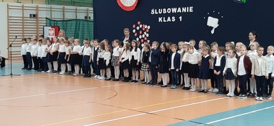 Uczniowie klas pierwszych ubrani w biało-granatowo stroje, z wychowawczyniami stoją na granatowym tle  z napisem ślubowanie klas pierwszych