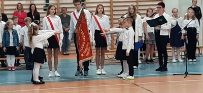 Sztandar szkoły, a przy nim dzieci strojach ślubujące z wyciągniętymi prawymi rękoma .
