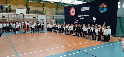 Dzieci ubrane w biało-czarne stroje z uniesionymi dwoma palcami składają przyrzeczenie na ucznia.