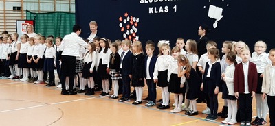 Uczniowie klas pierwszych ubrani w biało - granatowe stroje. W  tle granatowe płótno z dekoracją z kwiatów  oraz napis ślubowanie klas pierwszych. Pani dyrektor dokonuje pasowania na uczniów.