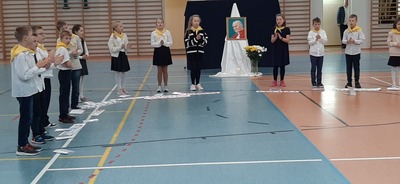 Uczniowie ubrani w odświętne stroje z chustami wokół szyi stoją w półokręgu. W tle portret Jana Pawła II 