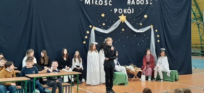 Uczniowie siedzą w ławkach a anioły stojące, Maryja, Józef i śpiewają, chłopiec śpiewa na tle szopki bożonarodzeniowej.
