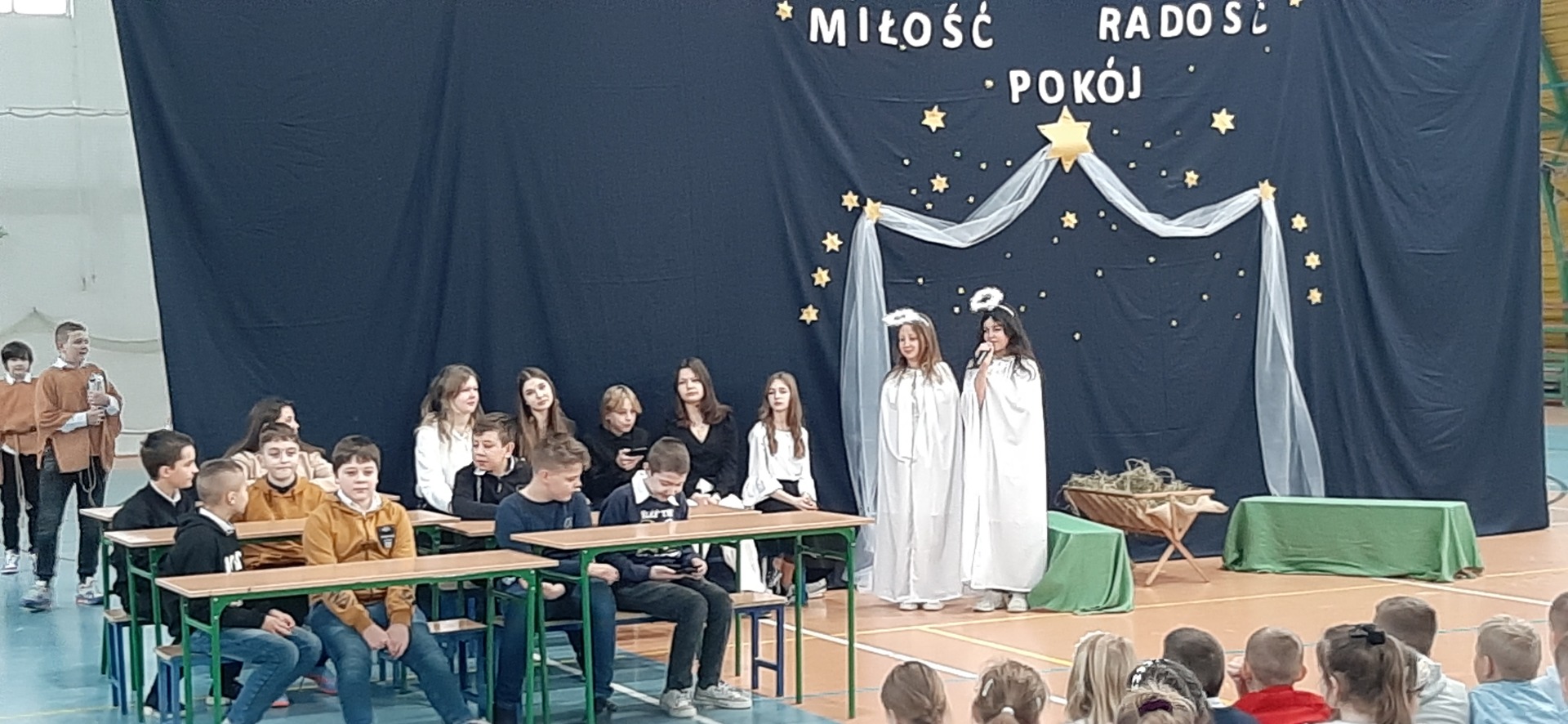 Uczniowie siedzą w ławkach a dziewczynki przebrane za anioły stoją na tle szopki bożonarodzeniowej.