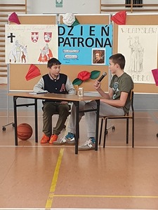 Dwóch uczniów siedzi przy stoliku na tle tablicy okolicznościowej.