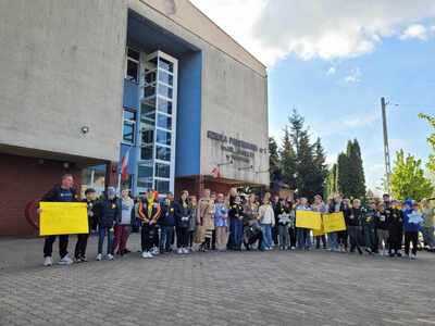Uczniowie stoją przed szkołą prezentując gotowość do marszu oraz plakaty z nazwiskami żydowskich uczniów naszej szkoły z 1926 roku.