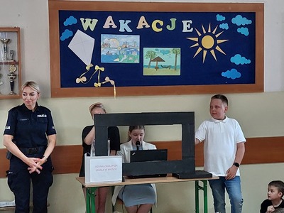 Troje uczniów biorących udział w apelu. Na ścianie wisi tablica z wakacyjną dekoracją. Obok uczniów stoi pani policjantka. 
