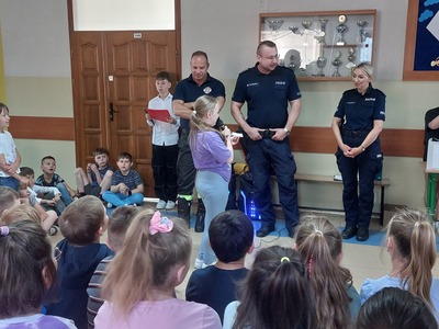 Grupa uczniów siedząca na ławkach na korytarzu szkolnym. Uczennica czyta  z kartki pytanie skierowane do jednego z policjantów.