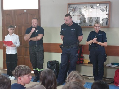  Korytarz szkolny. Grupa uczniów słuchająca wskazówek na temat bezpiecznych wakacji udzielanych przez strażaka..