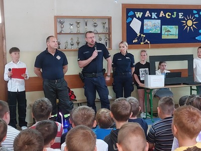  Korytarz szkolny. Grupa uczniów słuchająca wskazówek na temat bezpiecznych wakacji udzielanych przez policjanta