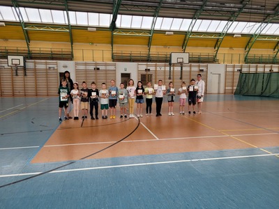  Hala sportowa. Grupa uczniów z klas drugich stoi w szeregu, trzymając dyplomy. Obok nich stoi pani nauczycielka oraz dyrekcja szkoły.