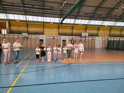 Hala sportowa. Grupa uczniów z klas pierwszych stoi w szeregu, trzymając dyplomy. Obok nich stoją nauczycielki i dyrekcja szkoły.