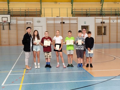 Hala sportowa. Grupa  uczniów z klasy 5b stoi w szeregu, trzymając dyplomy. Obok nich stoi wychowawczyni  i dyrekcja szkoły.