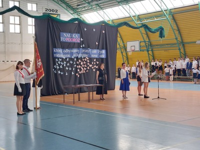 Hala sportowa. Na środku stoi Dyrekcja szkoły, obok stoi Sztandar Szkoły. Na ławkach siedzą uczniowie. W tle widać dekorację z okazji Zakończenia Roku Szkolnego.
