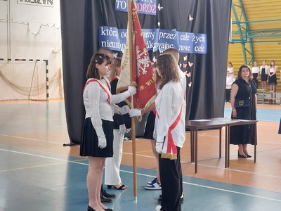 Hala sportowa. Uroczyste przekazanie Sztandaru Szkoły uczniom z klas siódmych. W tle stoi Pani Wicedyrektor oraz uczniowie.
