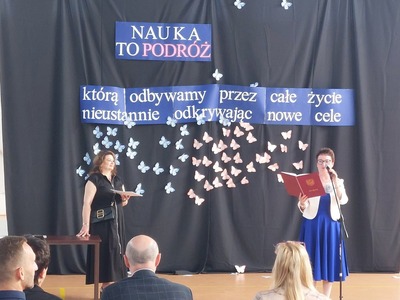 Hala Sportowa. Na tle dekoracji z okazji Uroczystego Zakończenia Roku Szkolnego przemawia do społeczności szkolnej Pani Dyrektor, a za nią stoi Pani Wicedyrektor.