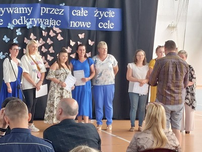 Hala Sportowa. Na tle dekoracji z okazji Uroczystego Zakończenia Roku Szkolnego stoją w rzędzie od lewej Dyrekcja szkoły. wyróżnieni Rodzice wraz z Wychowawczyniami uczniów z klas trzecich. 