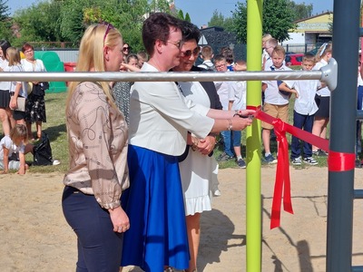 Teren zielony szkoły. Grupa osób - wśród nich m.in. zaproszeni goście, rodzice i uczniowie. Przy drabinkach do Street Workoutu Pani Dyrektor przecina wstęgę.