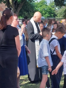 Teren zielony szkoły. Grupa osób - wśród nich m.in. uczniowie, ksiądz, Pani Dyrektor, która przemawia do społeczności szkolnej.