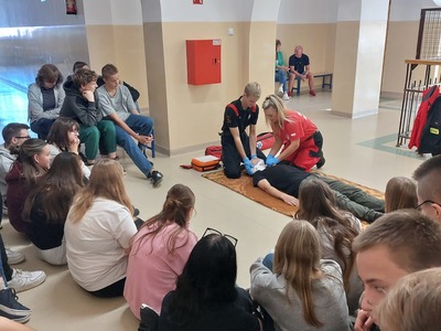 Strażak oraz ratownik medyczny prezentują na jednym z uczniów jak zabezpieczyć poszkodowanego  z podejrzeniem urazu kręgosłupa.