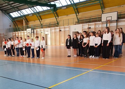 Boczny widok na uczniów ubranych w stroje galowe w hali sportowej  prezentujący się przed innymi uczniami.