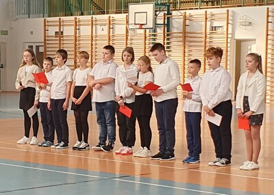 Zbliżenie na uczniów ubranych w stroje galowe, występujących  przez zgromadzonymi na hali sportowej.