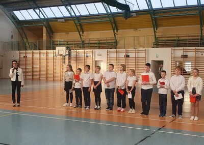 Przemawiająca pani dyrektor w otoczeniu uczniów na hali sportowej.
